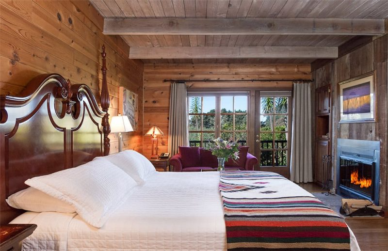 A cozy bedroom with wooden walls and ceiling, featuring a large bed with white linens, a striped blanket, and pillows. There are two bedside lamps, a small table, a purple sofa near large windows with view of trees, and a mounted fireplace with a fire burning.