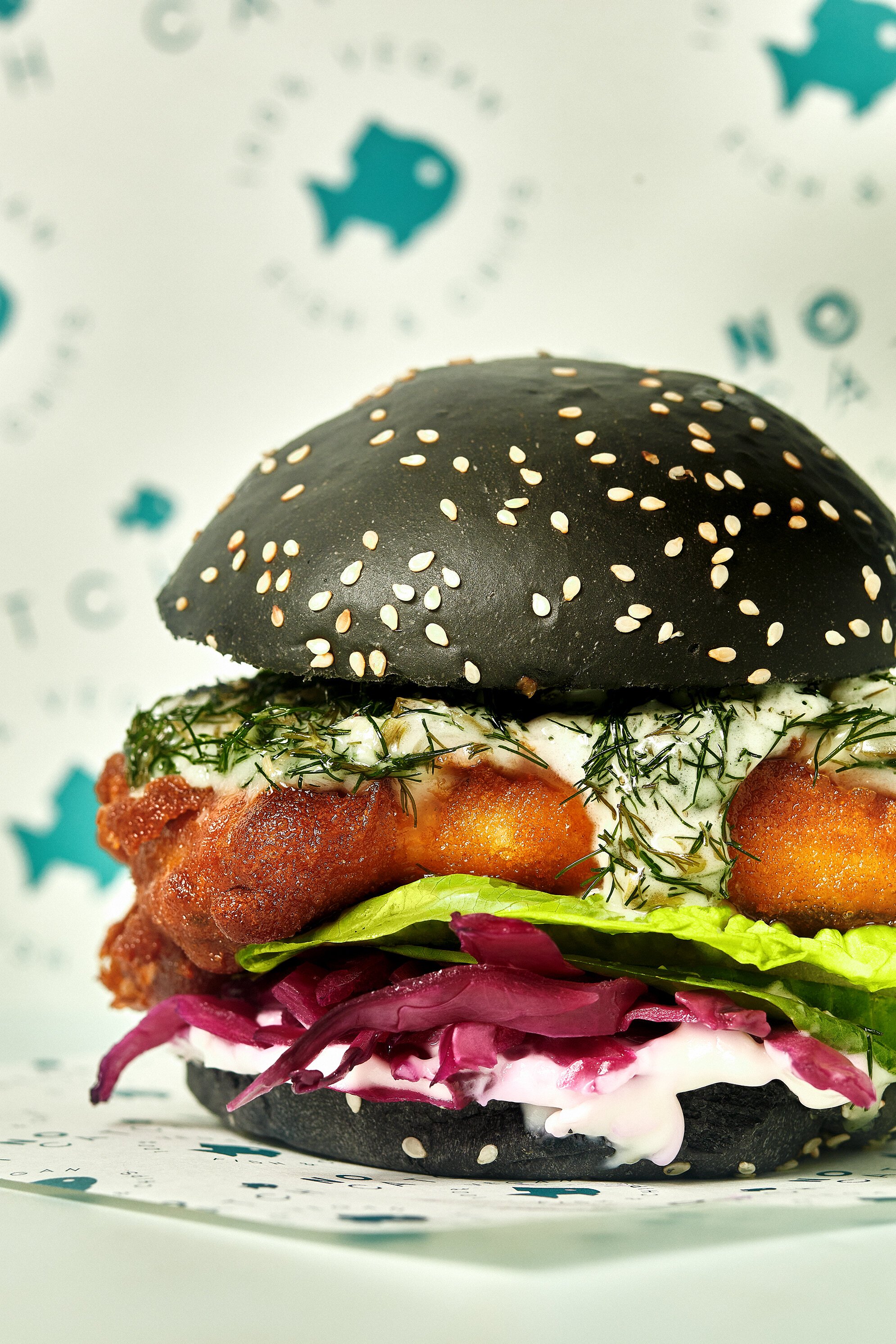 Close-up of a gourmet burger with black sesame bun, topped with white sesame seeds, lettuce, a tomato slice, herbs, and sauce, on a decorative paper sheet with fish and starfish illustrations.