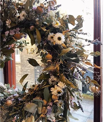 A floral arrangement with white daisies, yellow flowers, green leaves, and various other flowers and foliage on a window sill, with city buildings outside.