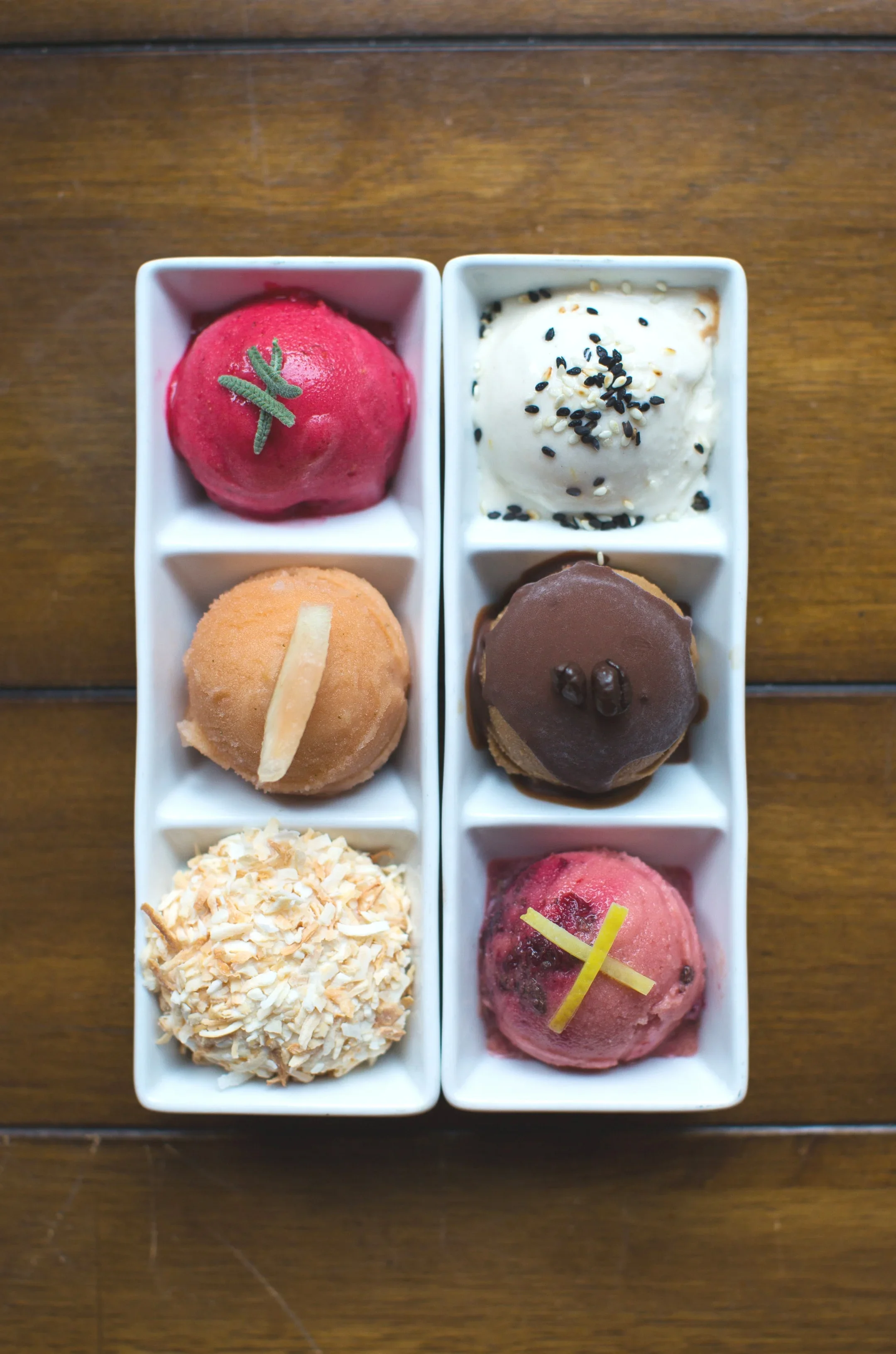 Six different scoops of ice cream in white rectangular dishes arranged on a wooden surface, with flavors including pink with green decoration, white topped with black and white sesame seeds, light brown with a white chocolate piece, dark brown with c