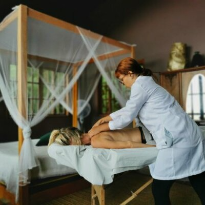 A massage therapist gives a back massage to a woman lying on a massage table in a serene room with a four-poster bed and canopy.