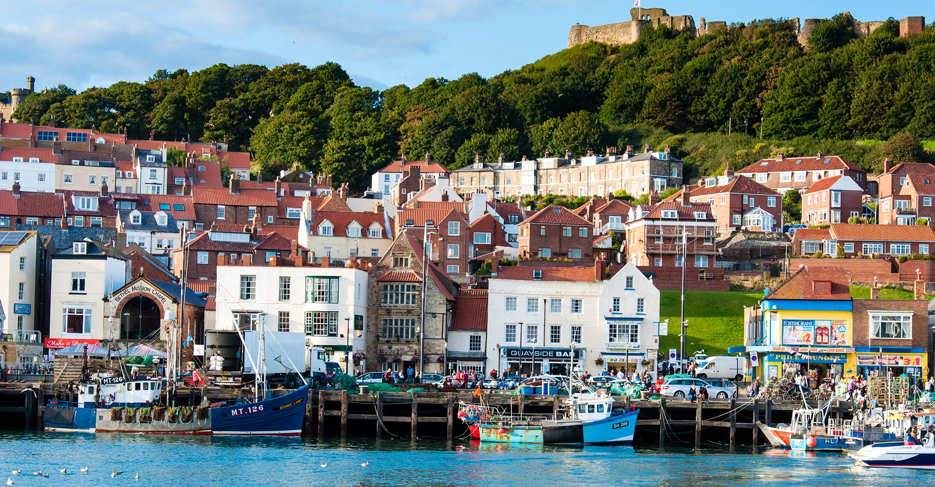 A harbor with boats docked and a promenade lined with shops and pubs set against a hillside with densely packed houses and a castle at the top.