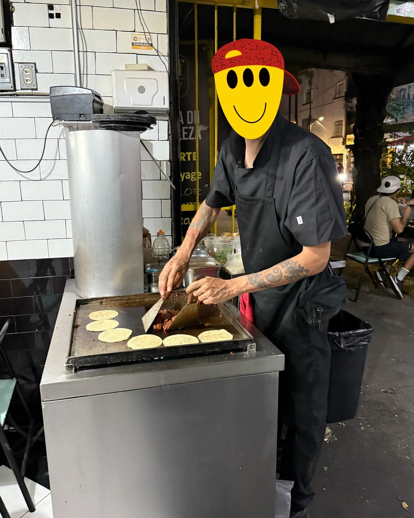 A street food vendor cooking tacos on a griddle, with tortillas and meat, at night outside a restaurant with people sitting nearby.
