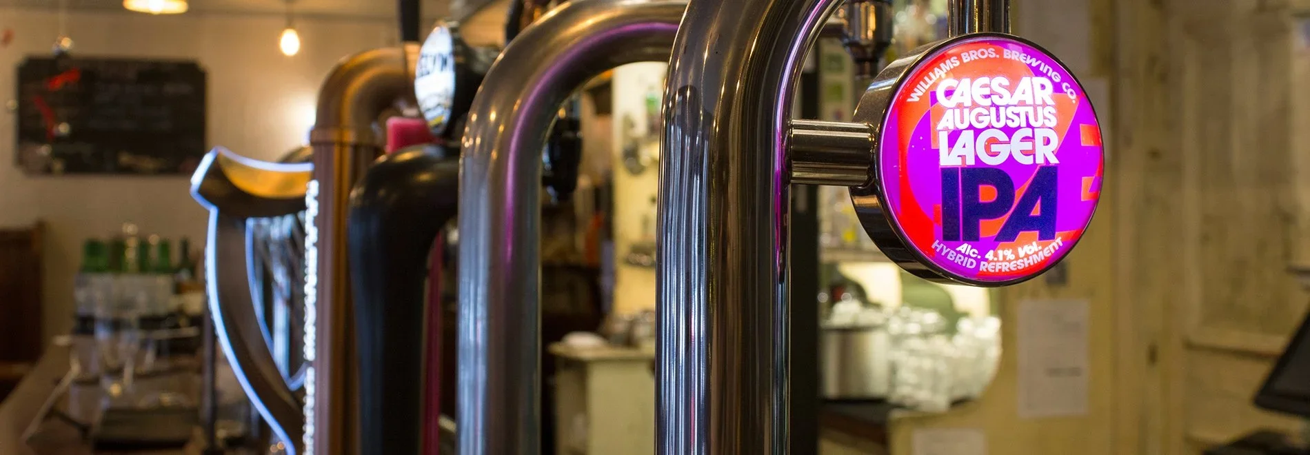 Close-up of beer taps with illuminated round sign showing Cesar Augustus Lager IPA, 4.1% alcohol, hybrid refreshment, on a bar counter.