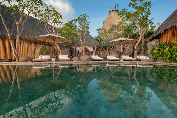 A tropical resort pool area with lounge chairs and umbrellas, surrounded by trees, thatched-roof huts, and a thatched tower in the background.