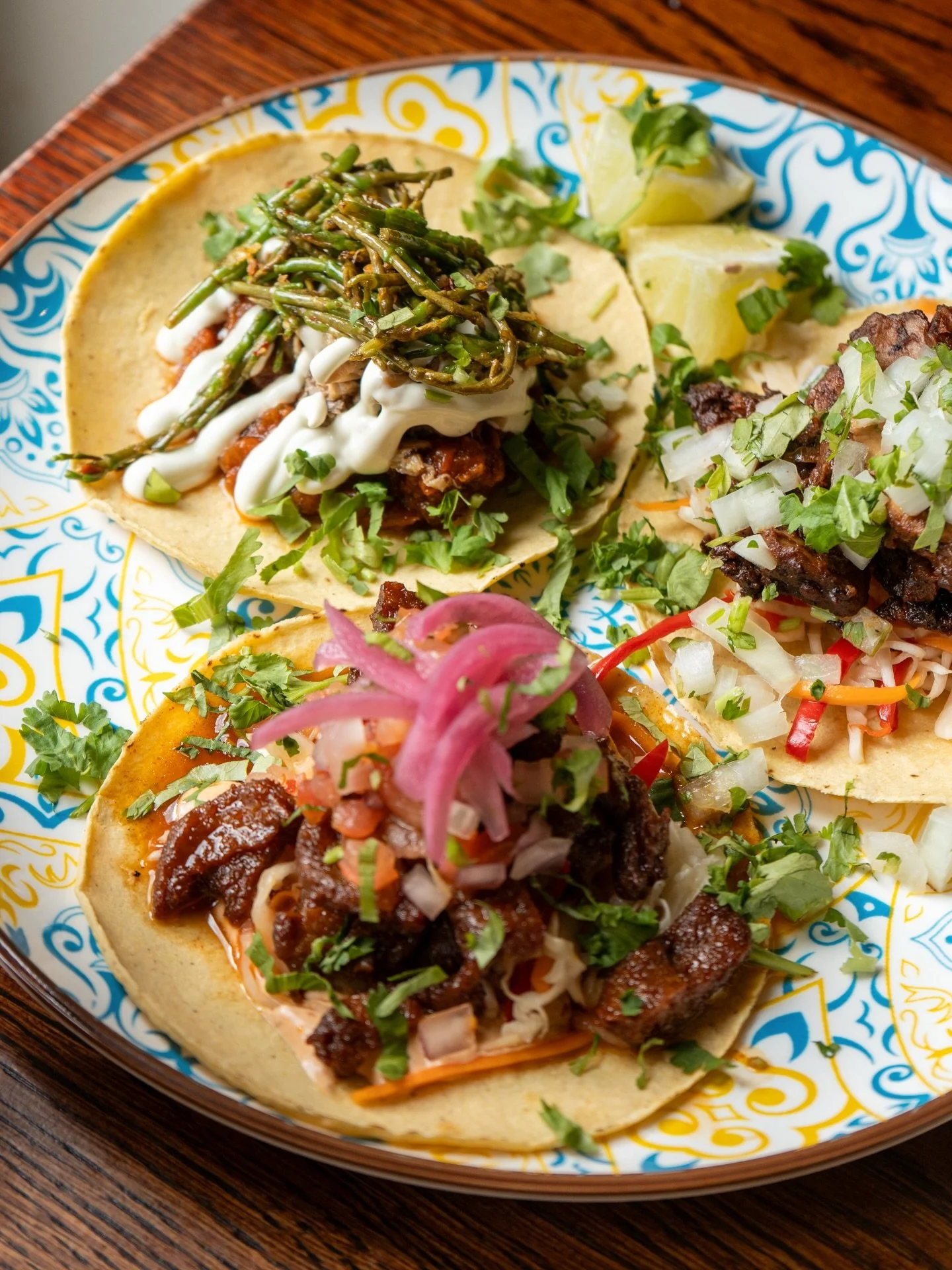 Four tacos on a decorative ceramic plate with colorful patterns, filled with meat, topped with sliced onions, cilantro, and garnished with chopped herbs, served on a wooden table.