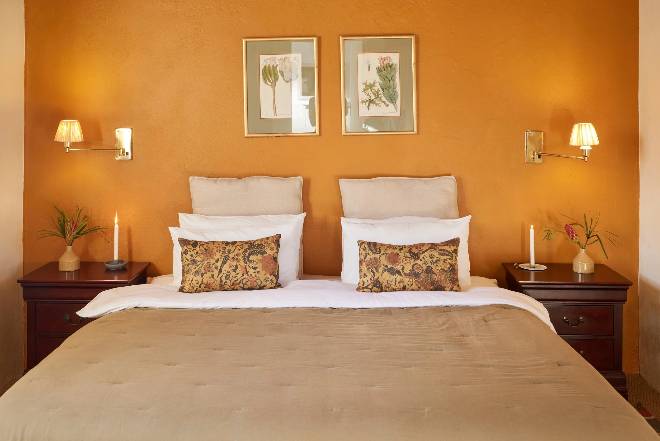 A neatly made bed with two white pillows, decorative pillows, and a beige quilt against an orange wall. There are two bedside tables with lamps, candles, and decorative plants. Above the bed are two framed botanical prints.