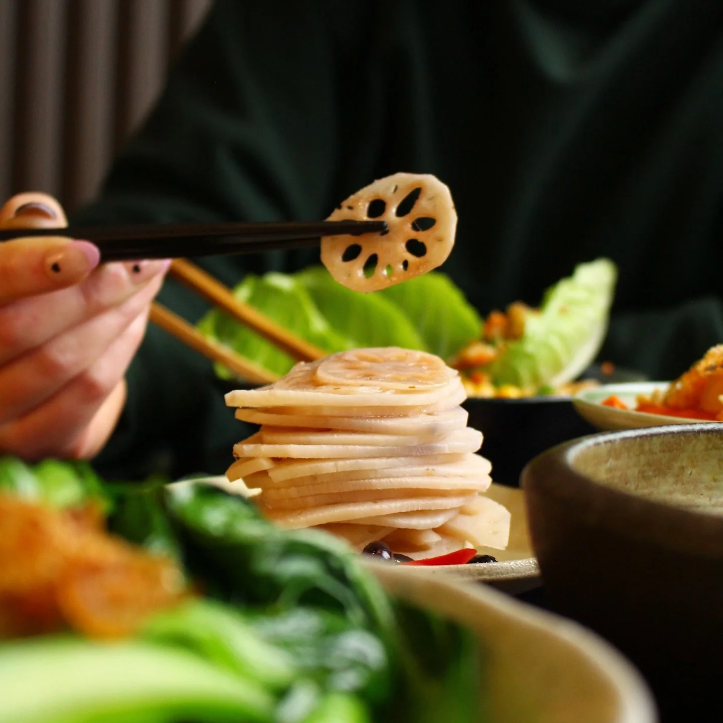 Close-up of a person's hand holding chopsticks with a slice of lotus root above a variety of Asian dishes on a table, including stacked sliced fish cake, leafy greens, and other colorful ingredients.