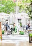 People sitting at outdoor tables in a park or café area, with umbrellas, trees, and greenery around.