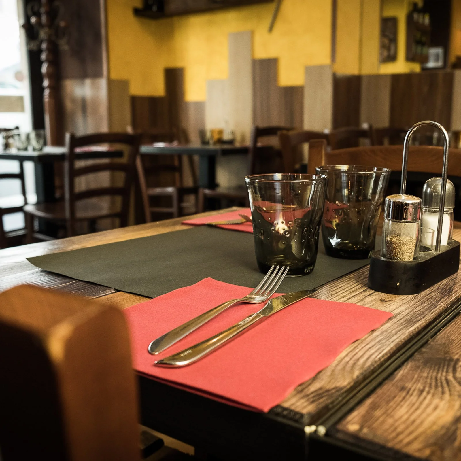 A close-up of a restaurant table setting with two empty glasses, a fork, a knife, and a red napkin on a black placemat, with wooden chairs and yellow-tan wall in the background.