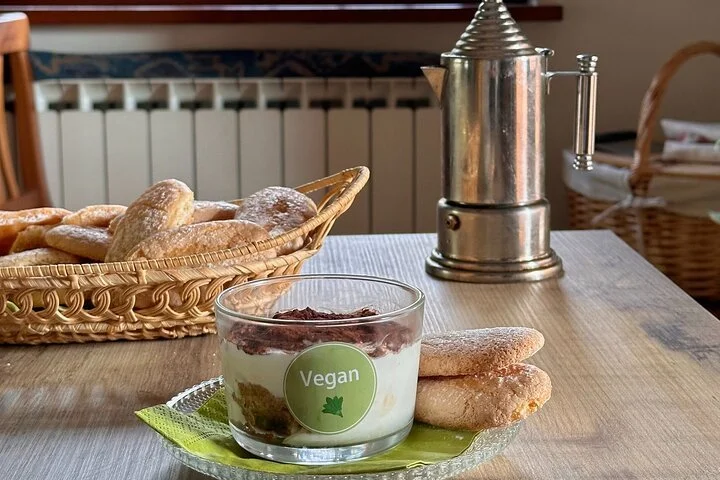 Vegan dessert in a glass bowl topped with a green sticker labeled "Vegan," served on a green plate with two cookies, on a wooden table with a basket of bread and a metal coffee grinder in the background.