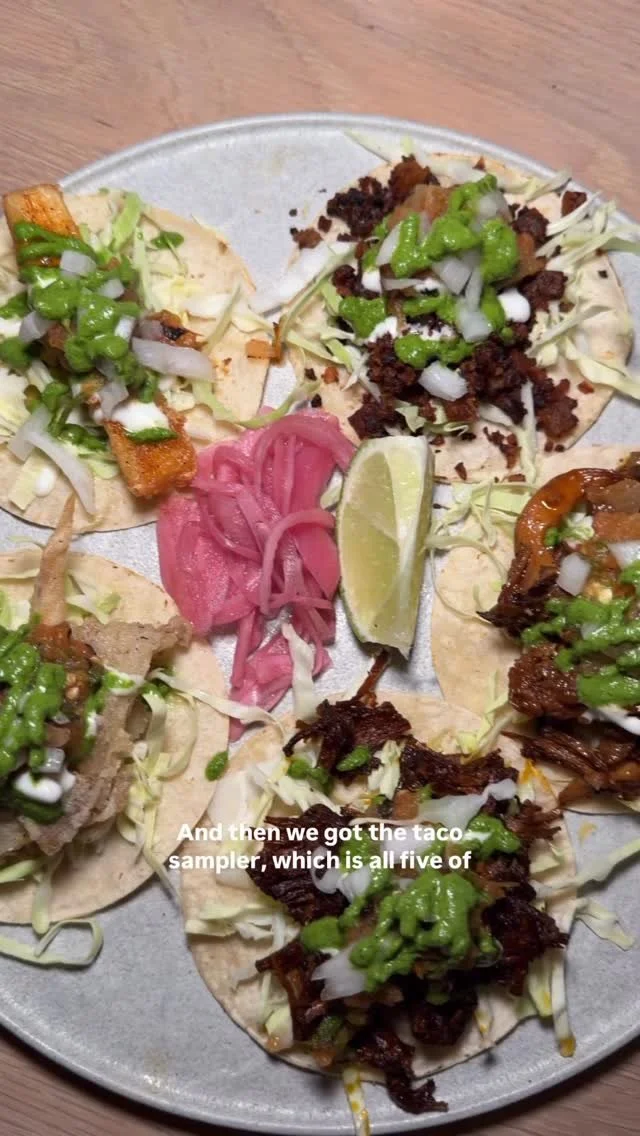 Assorted tacos with shredded lettuce, chopped onions, green sauce, and a lime wedge on a metal plate, served with pickled pink onions.