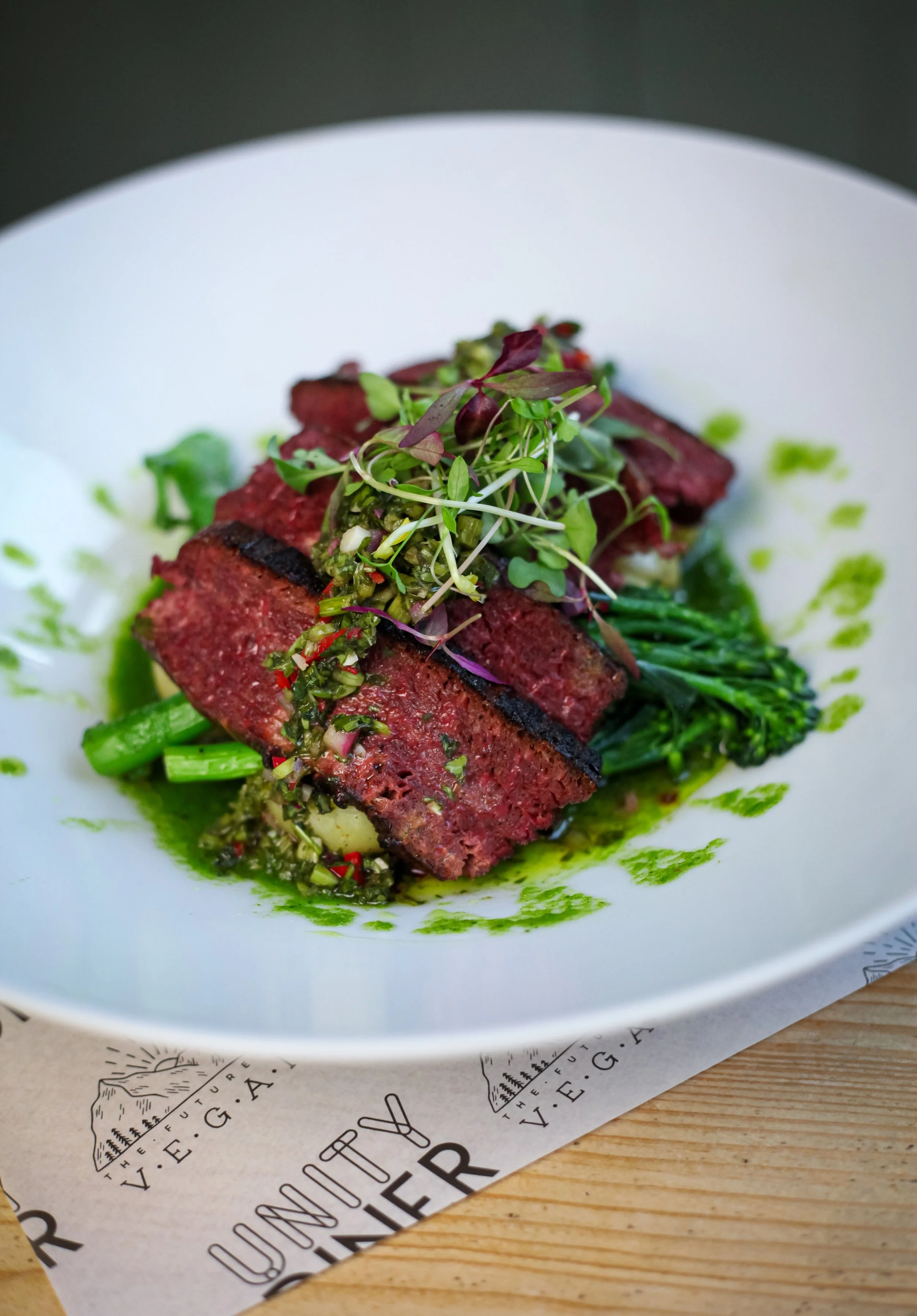 A plated dish with a piece of cooked meat topped with microgreens and a green sauce, served with green vegetables on a white plate.