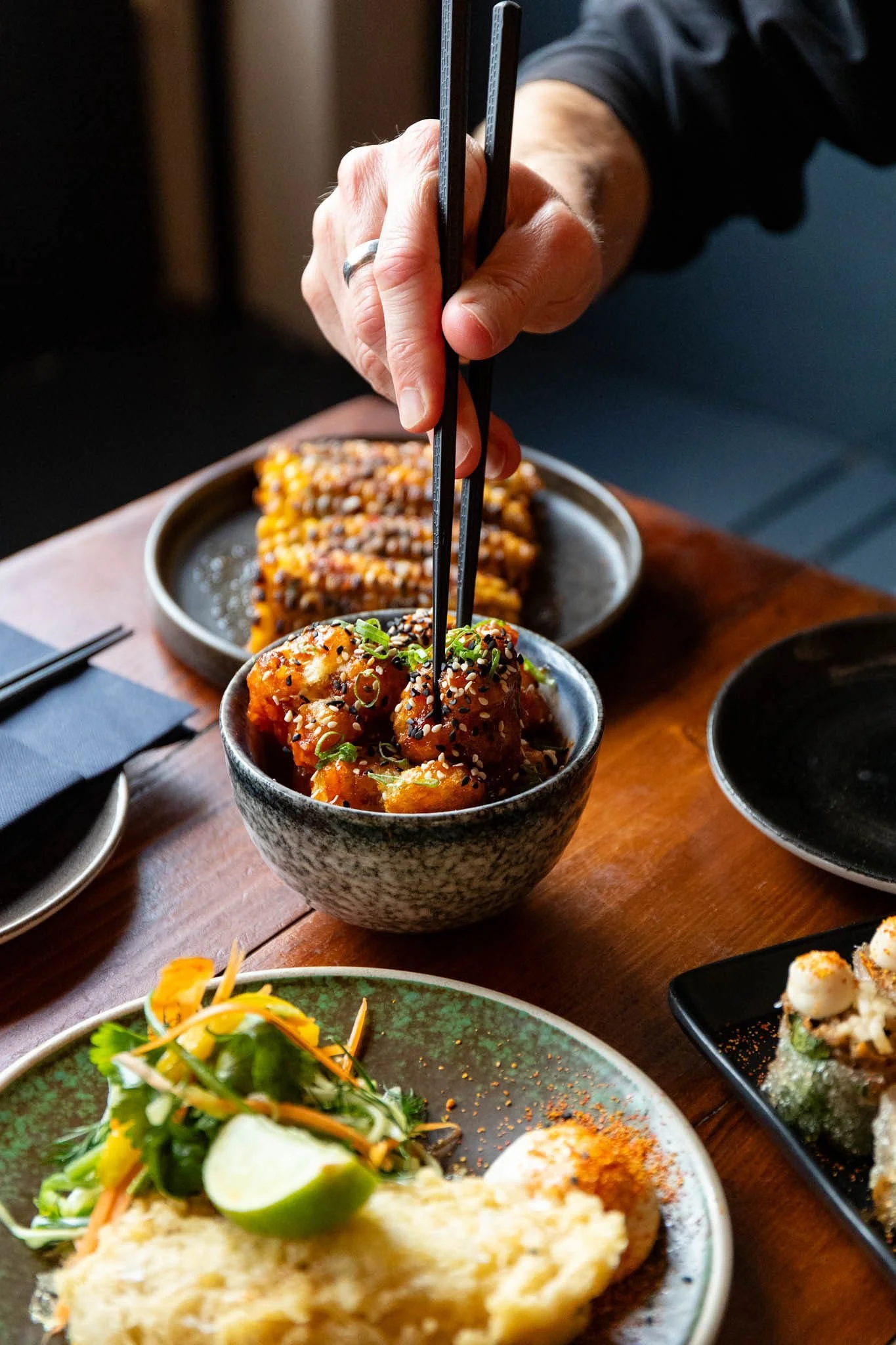 Person using chopsticks to pick up food from a bowl of sweet and spicy sesame chicken, with other Asian dishes on a wooden table.