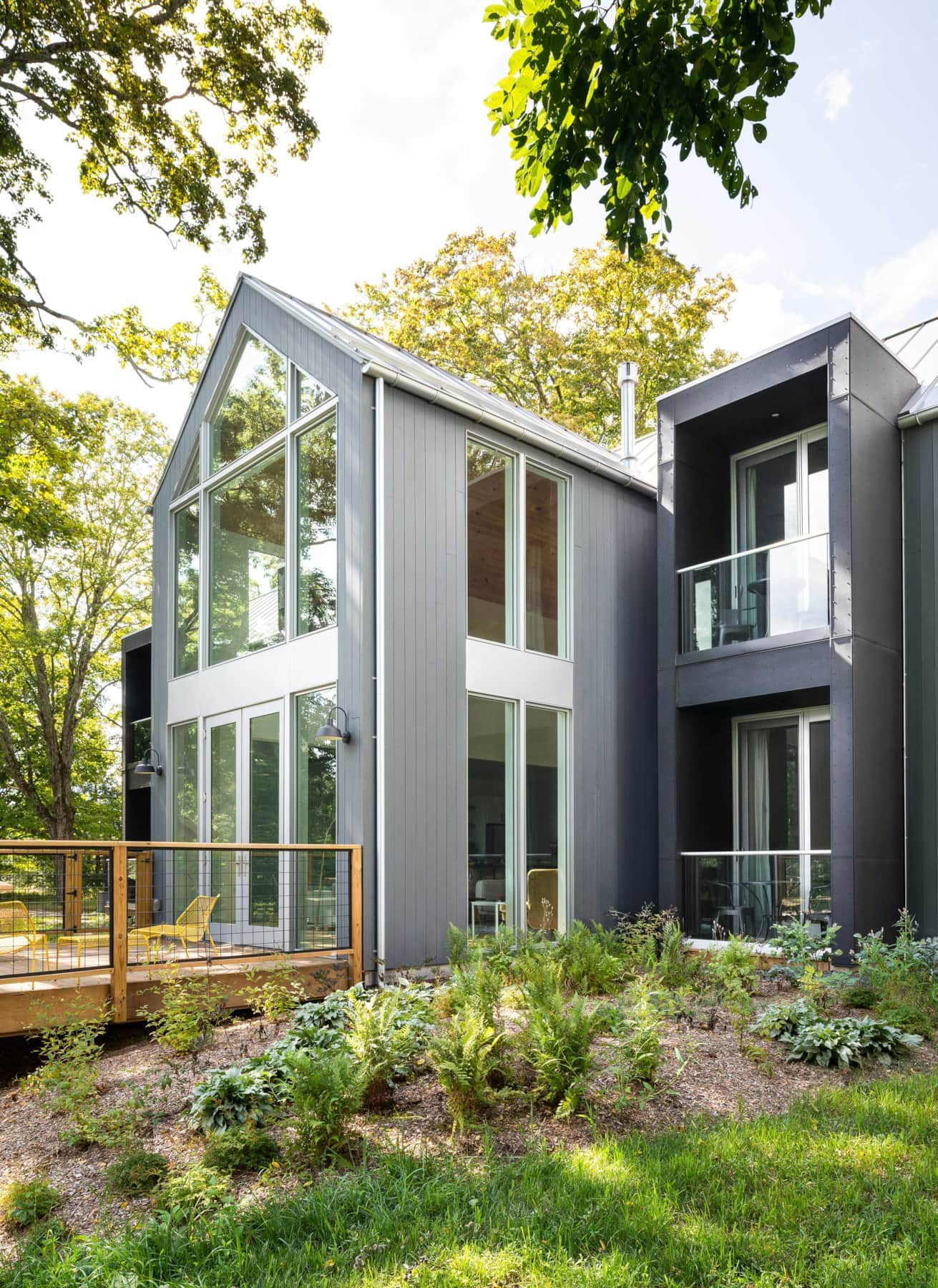 Modern gray multi-story house with large windows, balconies, surrounded by trees and greenery, with a small deck and patio furniture.