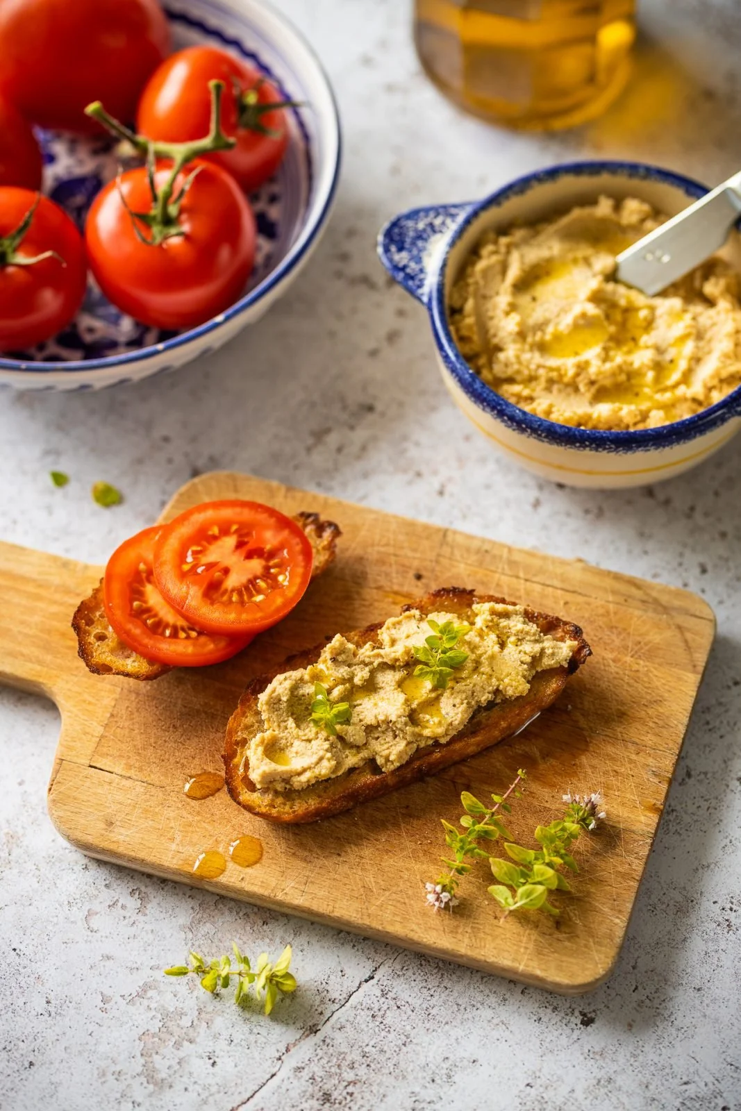 A wooden cutting board with a slice of toasted bread topped with hummus and garnished with herbs, next to two slices of tomato. In the background, there is a bowl of more hummus, a bowl of cherry tomatoes, and a glass of iced tea.