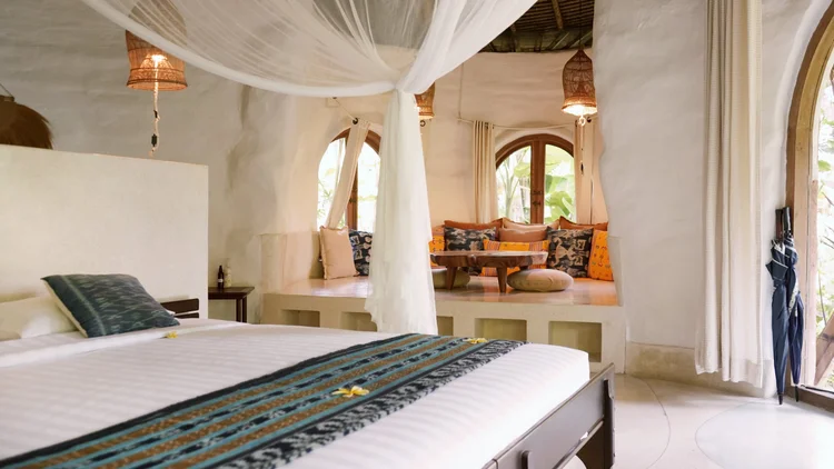 A cozy bedroom with a large bed, a seating nook with cushions by the windows, and hanging wicker lamps in a tropical-style room.