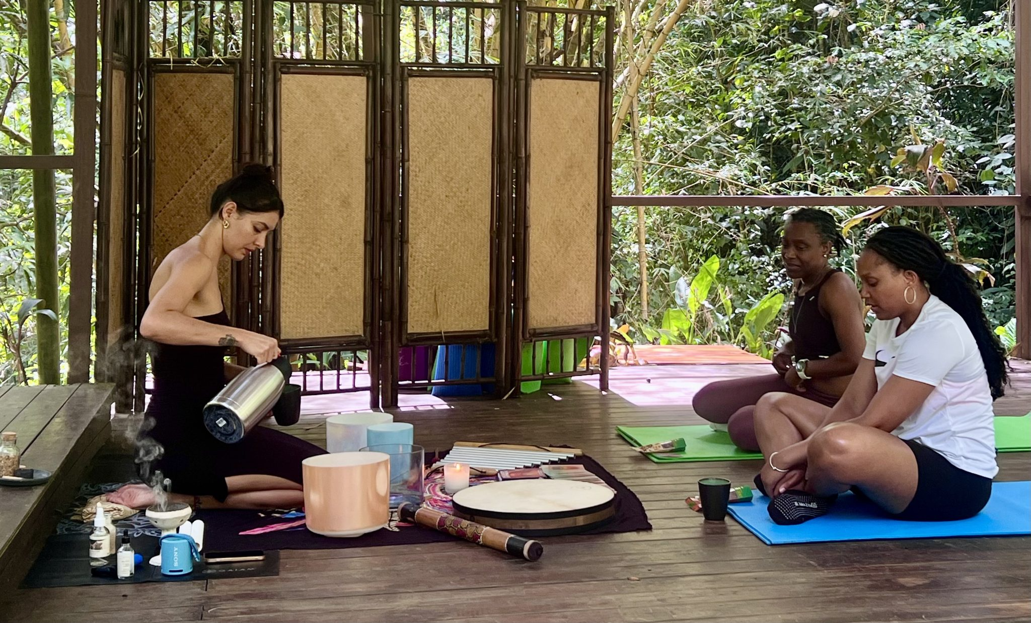 Three women participating in a yoga or meditation session inside a wooden, open-air structure surrounded by lush greenery. One woman is pouring a hot beverage, possibly tea, while the other two sit on yoga mats, with relaxed postures and closed eyes.