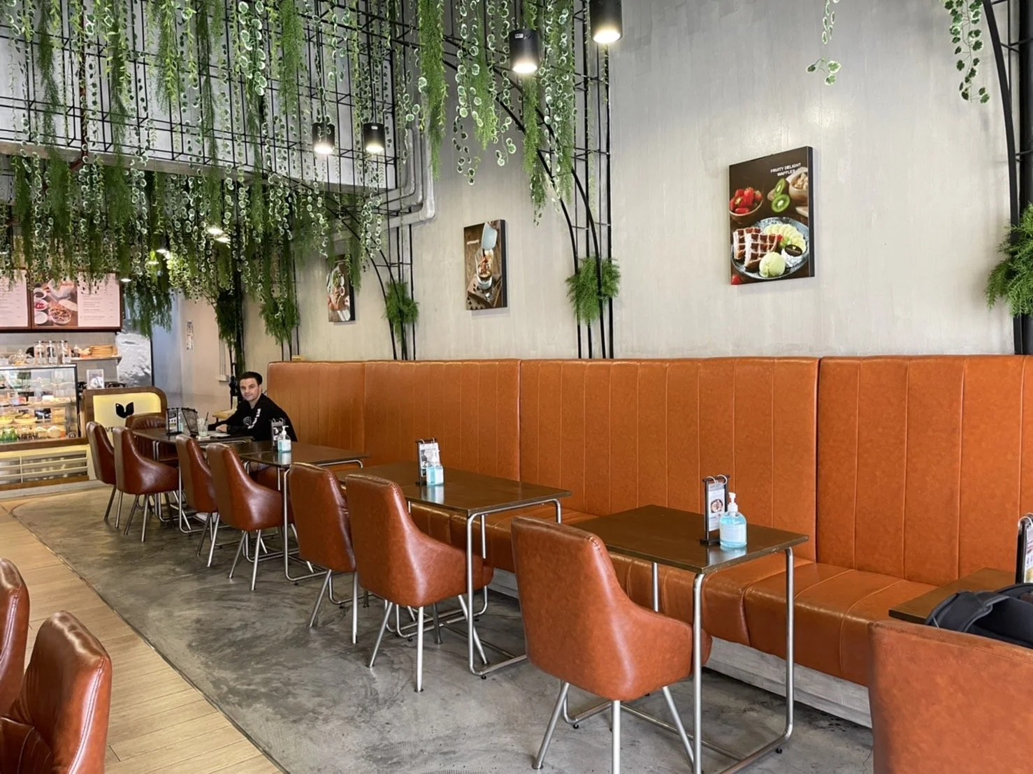 Interior of a modern cafe with a long orange upholstered bench, individual brown chairs, and small tables. Green hanging plants and framed food pictures decorate the light-colored wall. A person sits at one table, and a display case with baked goods 