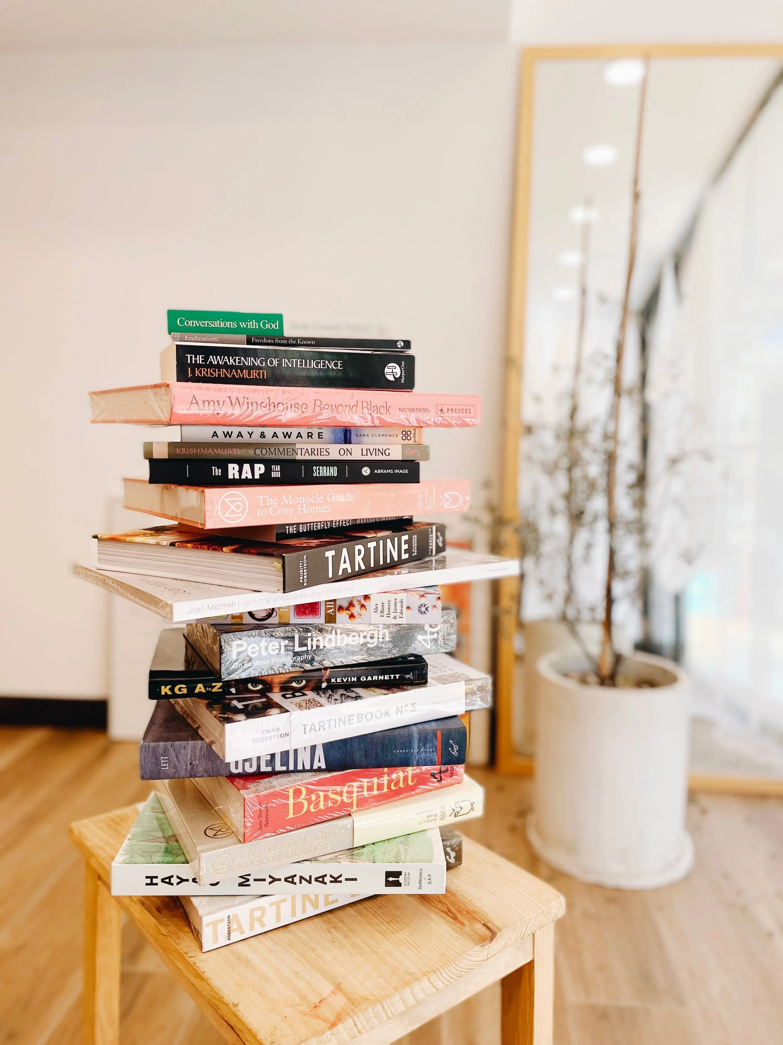 Stack of various books on a wooden table in a room with wooden floors and a potted plant in the background.