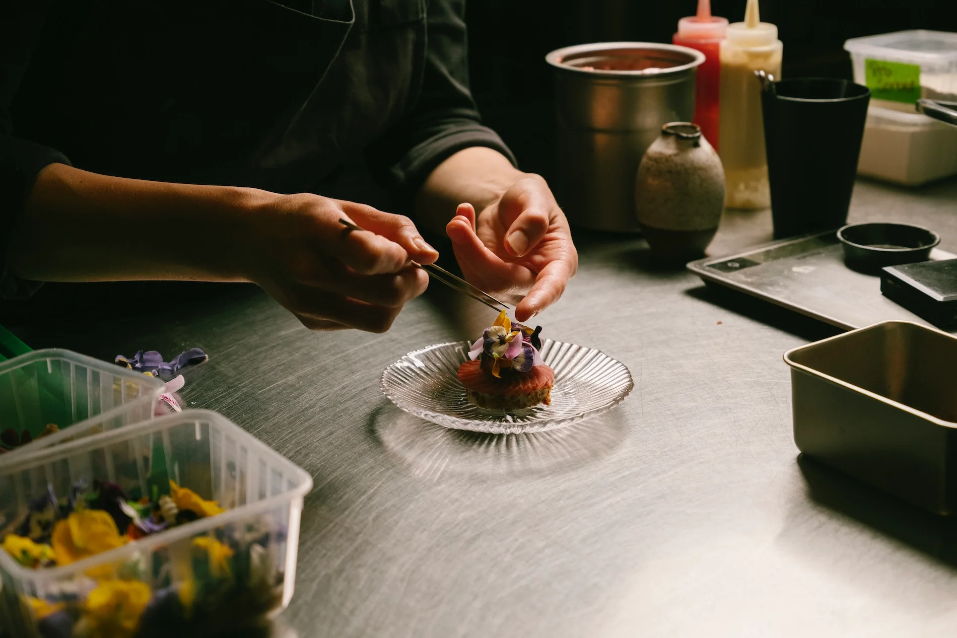 A chef decorates a dessert with edible flowers on a clear glass plate in a professional kitchen.