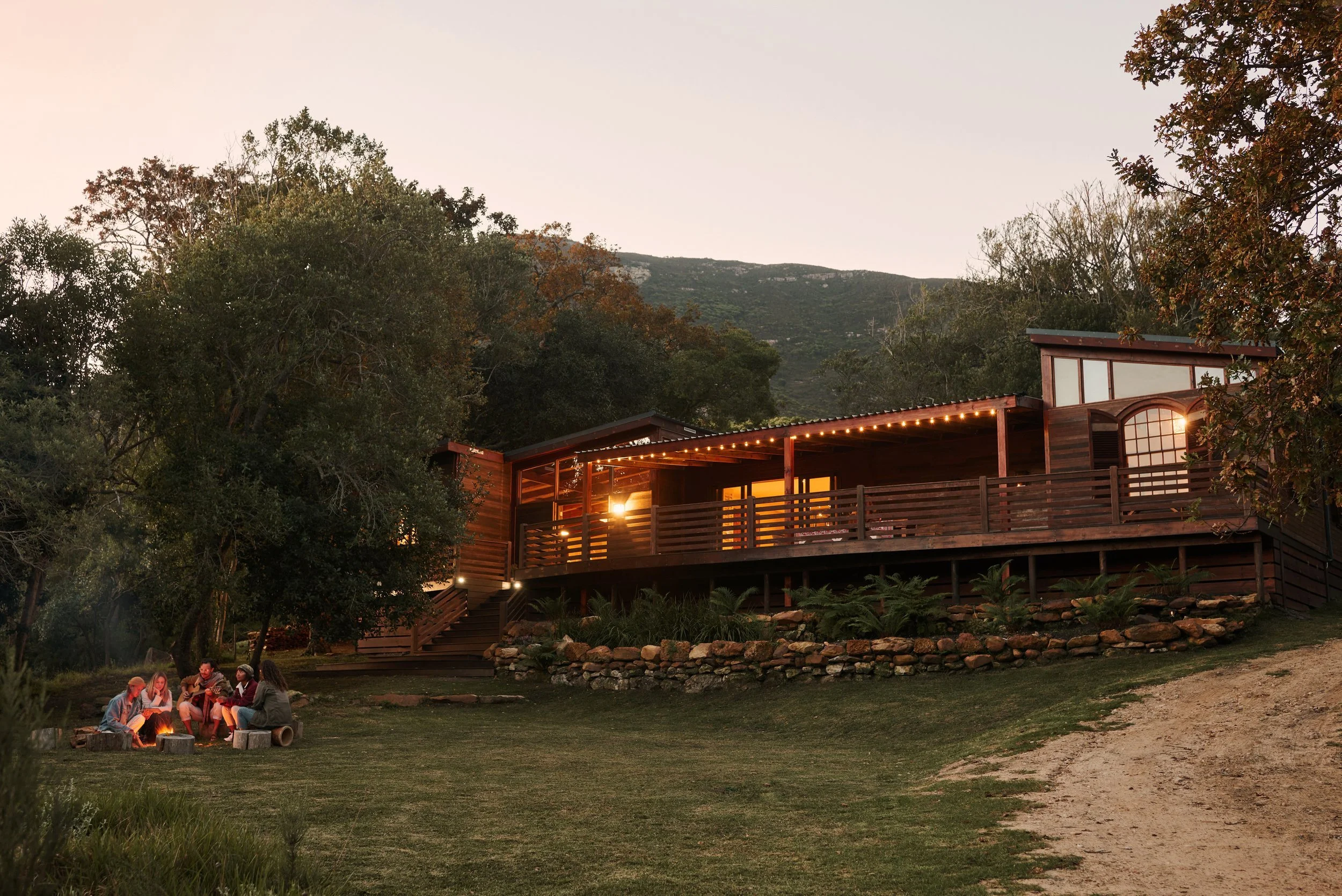 A group of people sitting around a campfire on a grassy area, with a hillside and trees in the background, and a modern house with string lights on the deck.