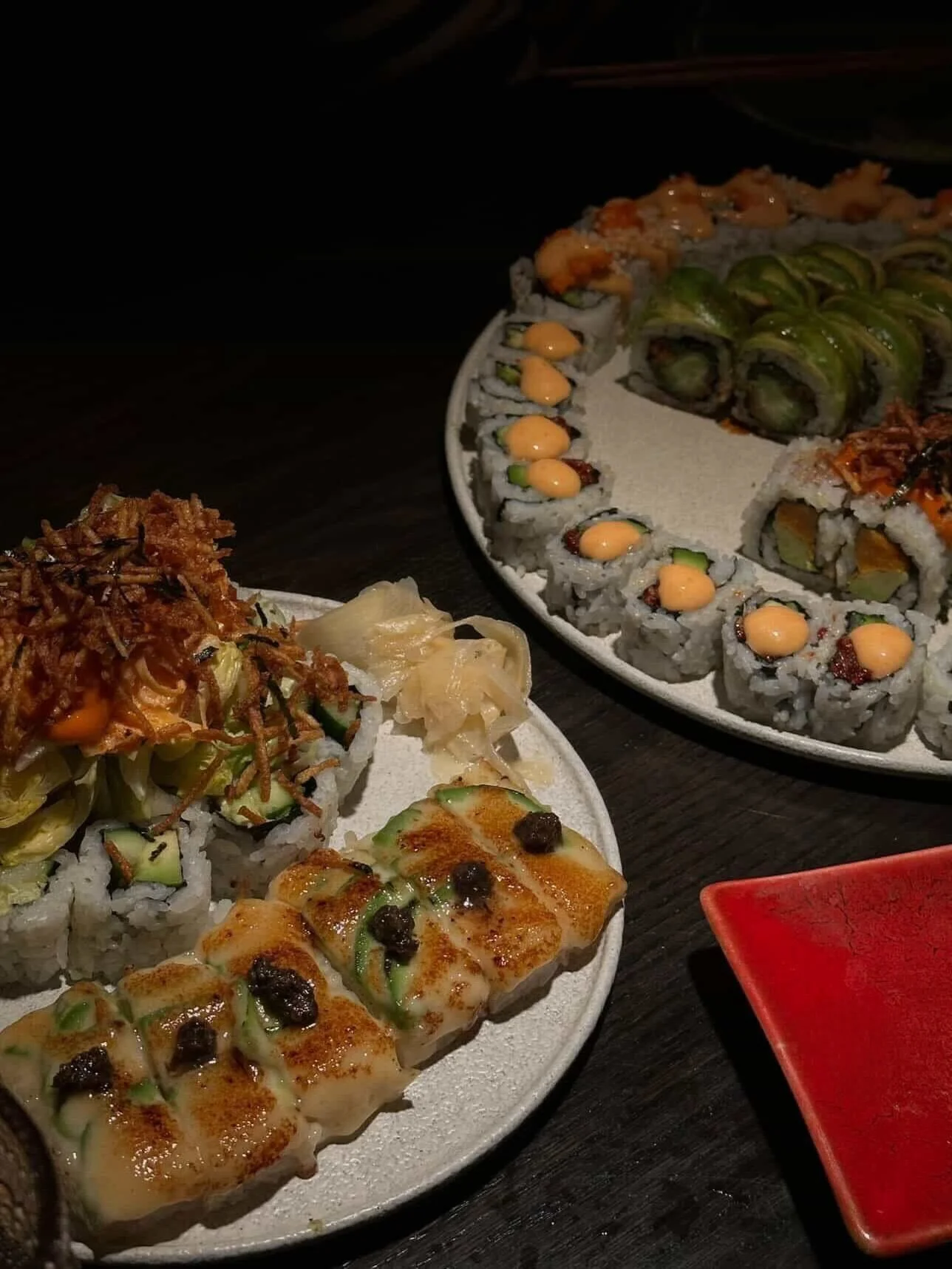 Assorted sushi rolls on white and black plates, served with soy sauce, pickled ginger, and wasabi on a dark table.