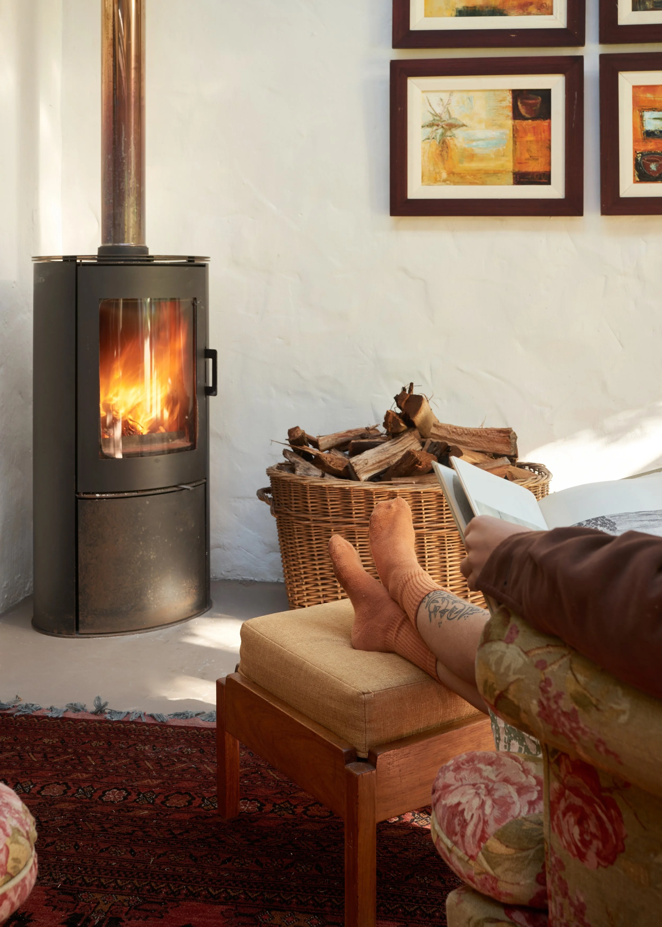 A person is relaxing on a sofa, wearing orange knitted socks, and reading a book. There is a wood-burning stove with a fire inside and a stack of firewood nearby. The room has white walls, framed abstract artwork, a patterned rug, and a wicker basket.