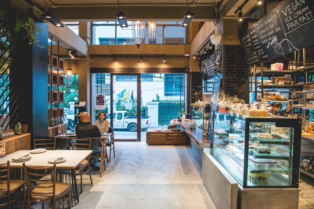 Interior of a cafe with seating, a display of baked goods, and a menu board, with large glass doors opening to the street outside.