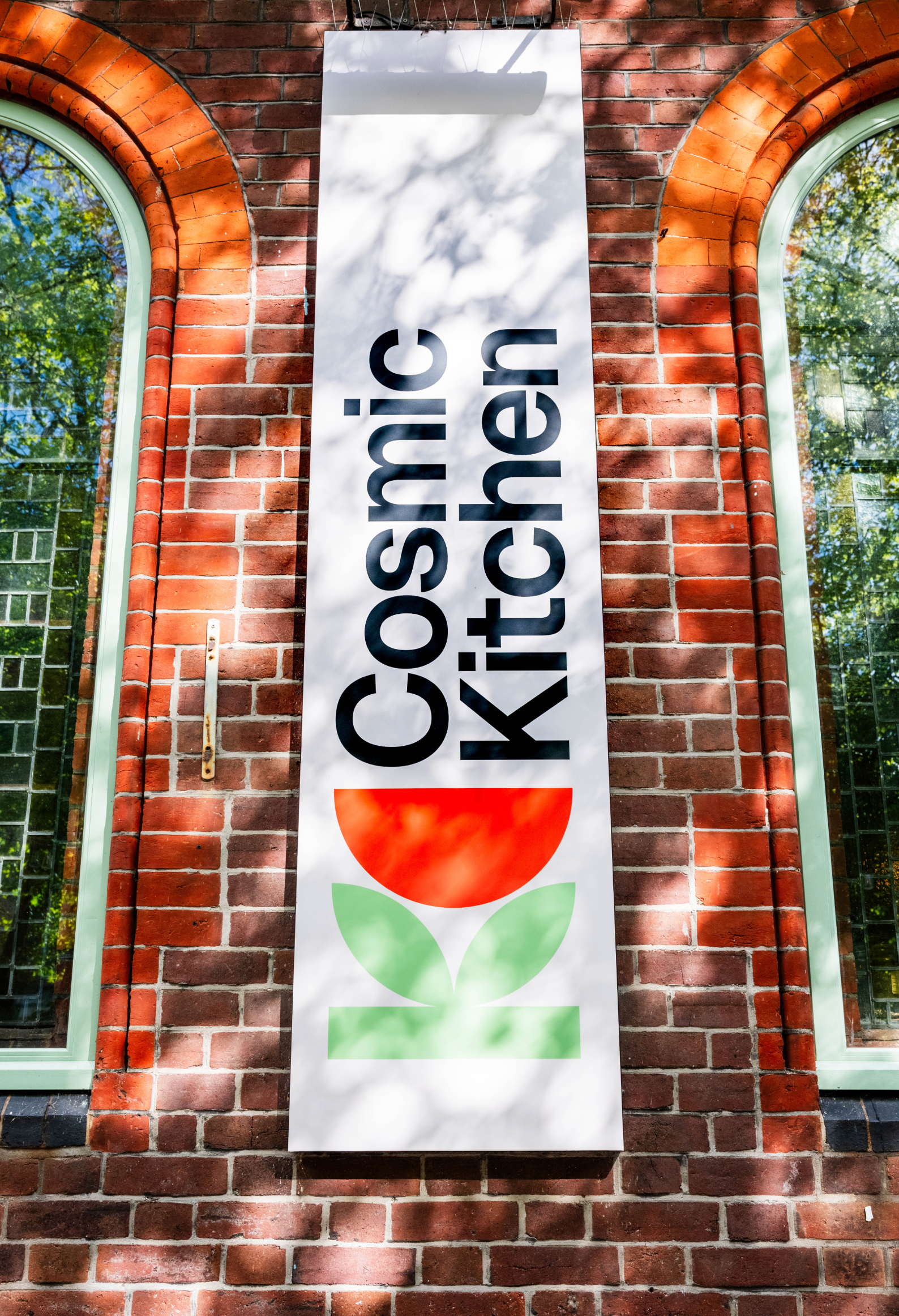 A vertical banner with the text 'CONSMİİ' and 'KİTAP EVI' in large black letters, hanging on a brick wall with two arched windows on either side, reflections of trees visible in the window panes.