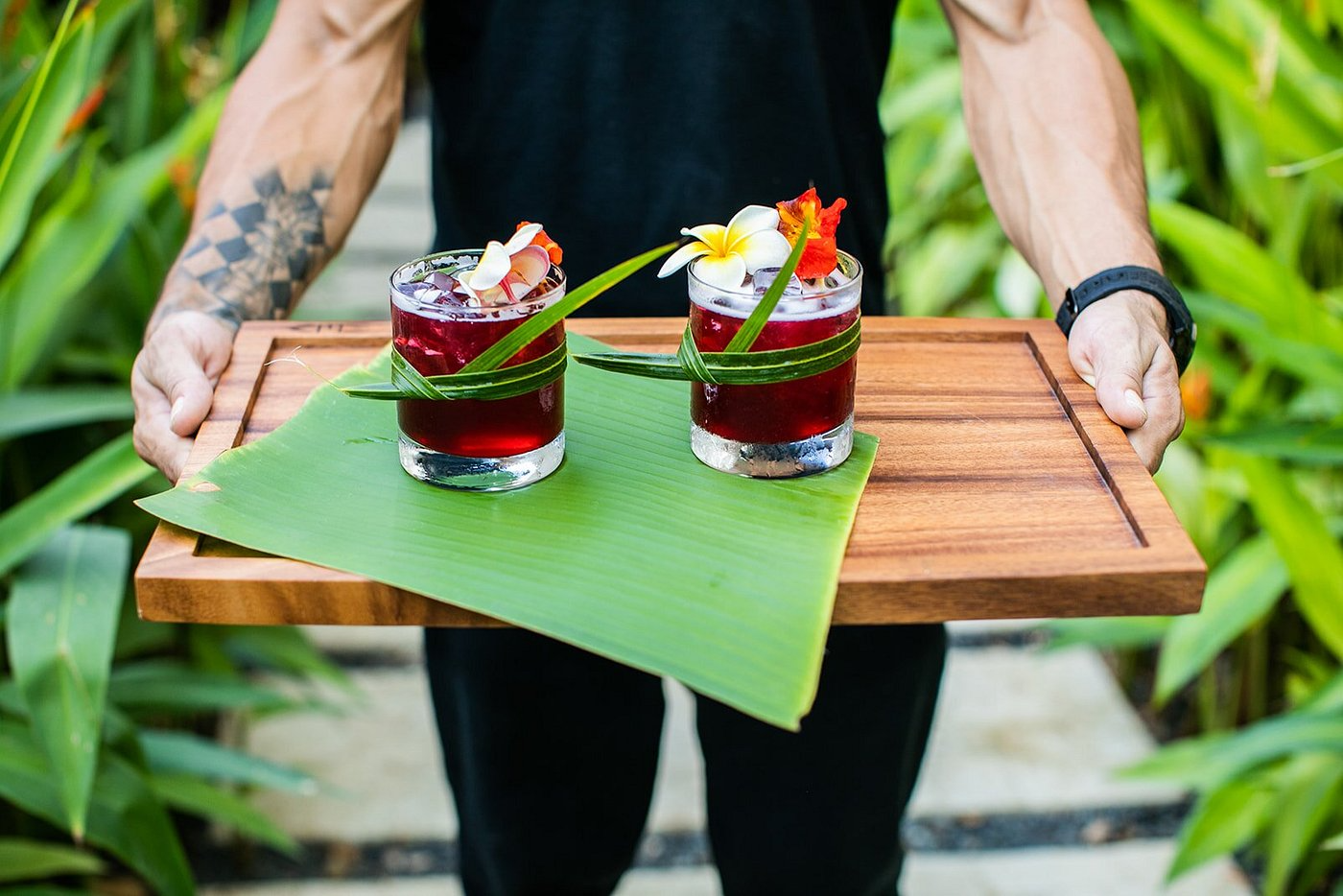 Two tropical drinks with red liquid, garnished with flowers and leaves, served on a green banana leaf on a wooden tray.