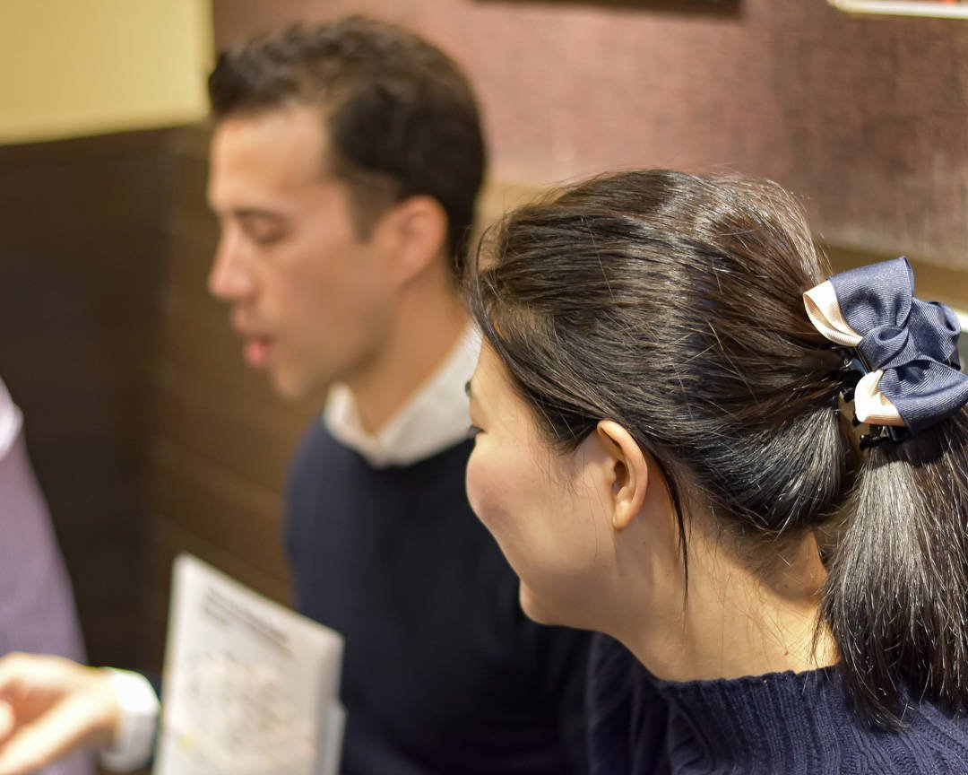 Woman with dark hair in ponytail wearing a bow hair accessory, smiling and talking to a man with short dark hair, indoors.