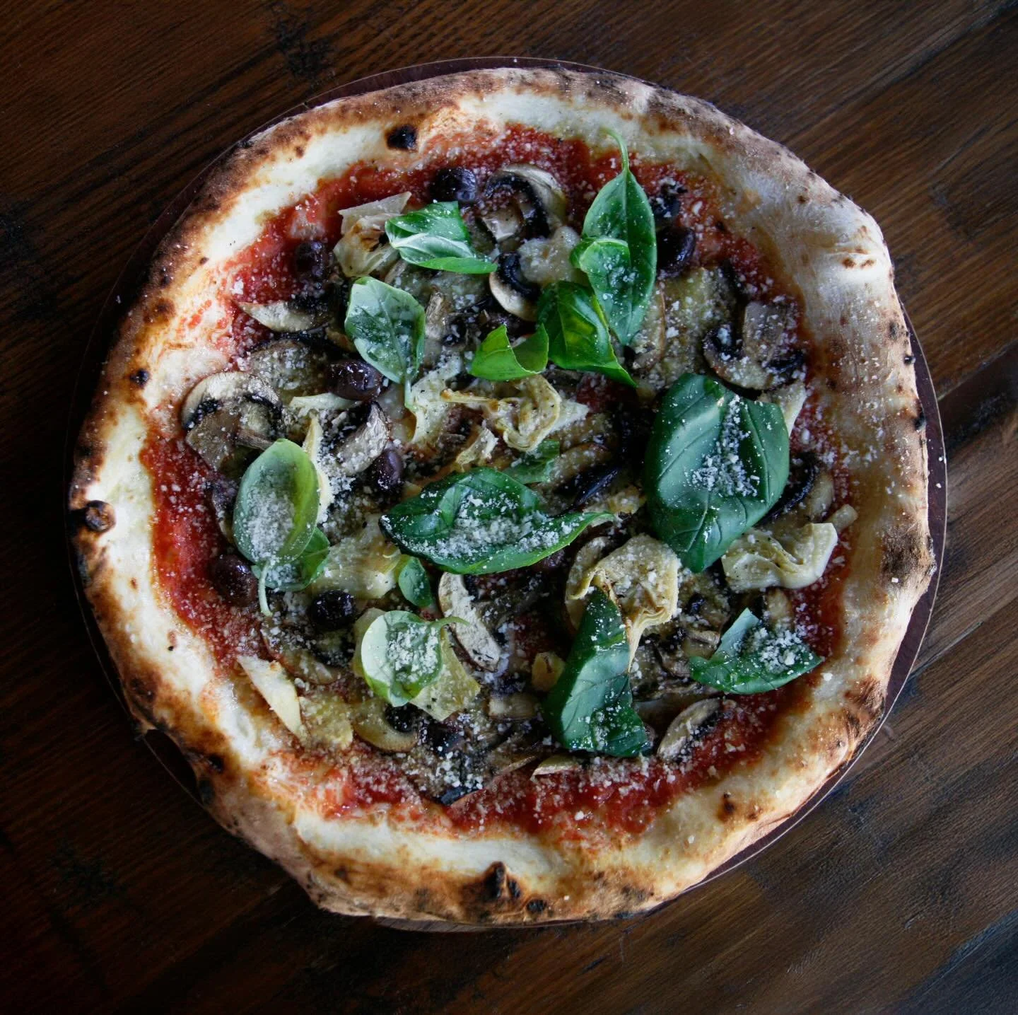 Top view of a freshly baked vegetable pizza with basil leaves, black olives, artichoke hearts, shredded cheese, and tomato sauce on a wooden table.