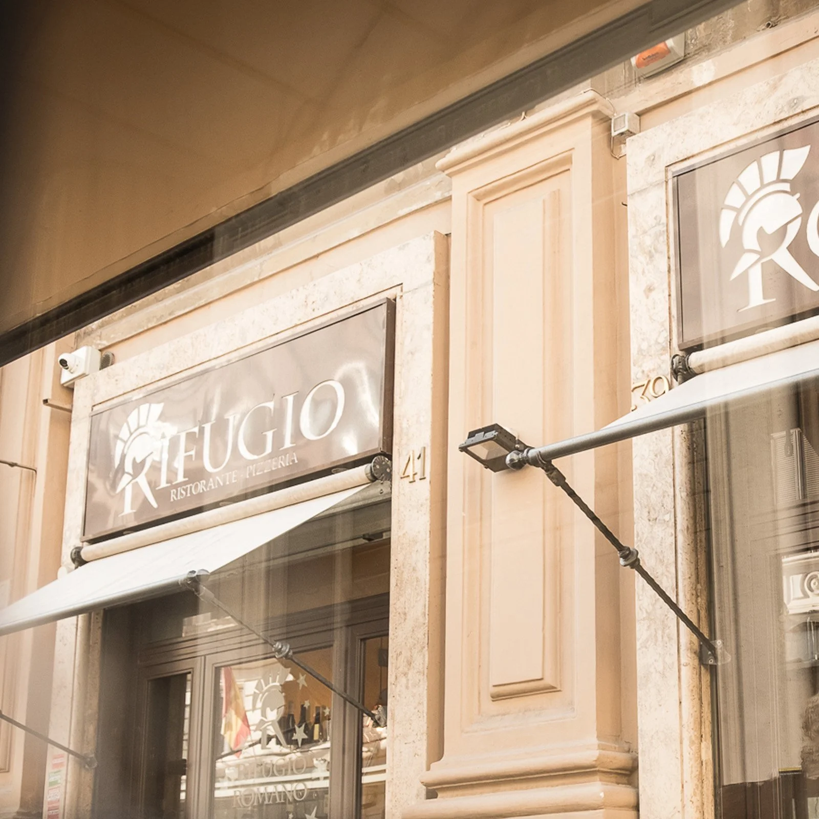 The image shows the exterior of a restaurant with a sign that reads 'FUGIO RISTORANTE - PIZZERIA.' There is a decorative beige-colored facade with stone accents and a security camera mounted near the upper left corner. A white awning extends over the