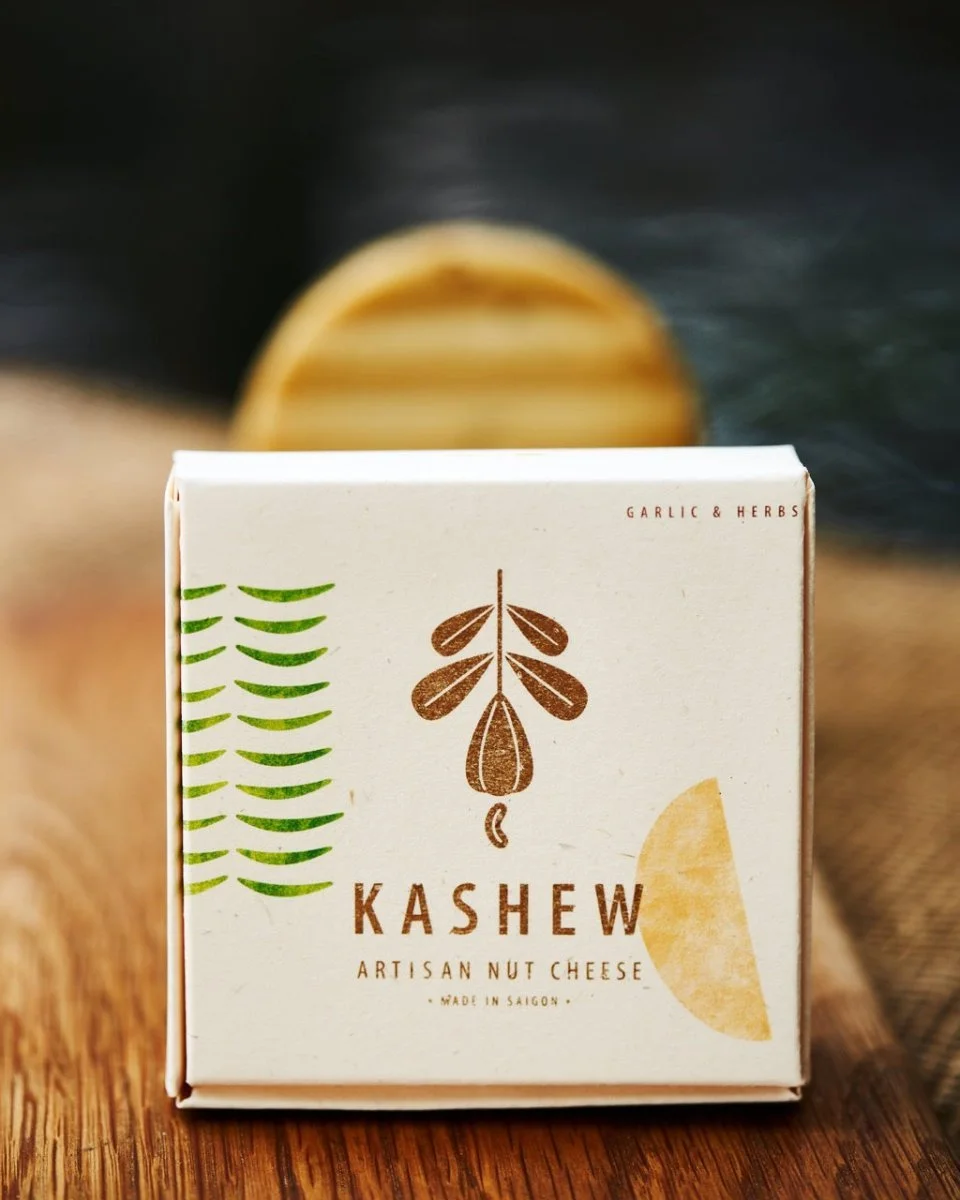 Front of a packaging box of Kashew artisan nut cheese, with a stylized plant illustration and text indicating garlic and herbs flavor, made in Saigon, placed on a wooden surface.
