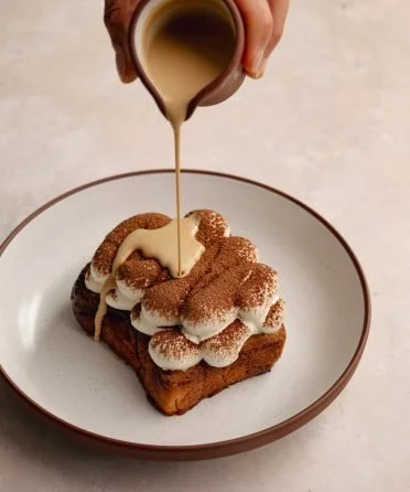 A slice of French toast on a white plate being topped with whipped cream and cocoa powder, with cream being poured from a small pitcher.
