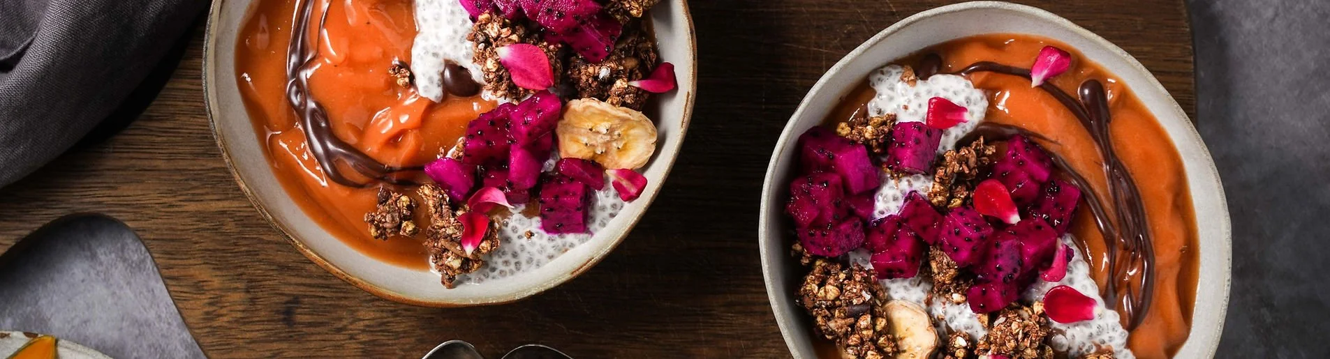 Two bowls of smoothie topped with dragon fruit, banana, granola, coconut, chia seeds, pink flower petals, and chocolate drizzle on a wooden table.