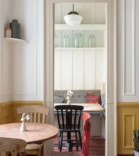 A cozy dining nook with a small round table, some chairs, and a built-in cushioned bench with pillows, separated from the main room by a narrow doorway. Decor includes glass jars on a shelf and a small vase with flowers on the table.