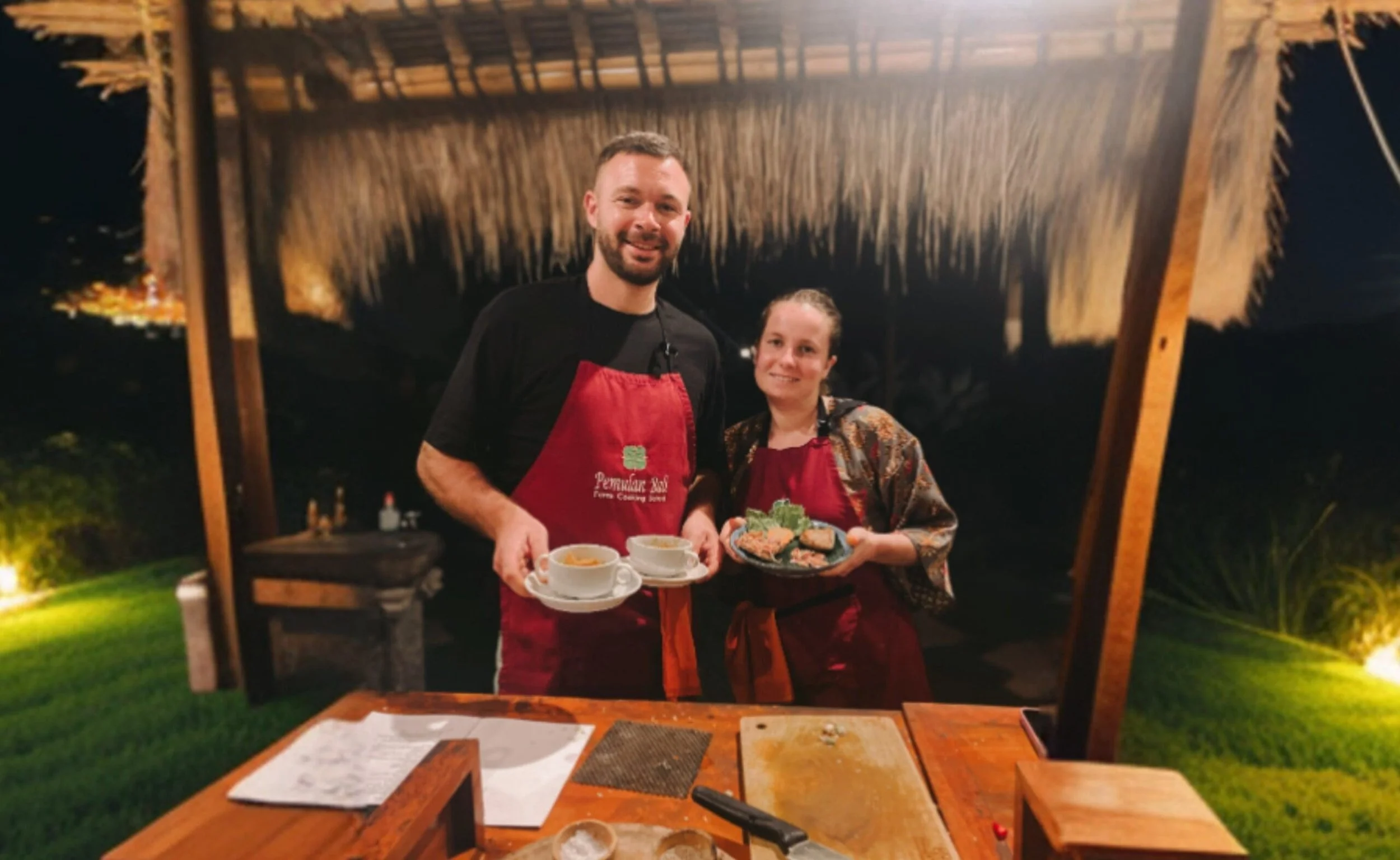 Two people, a man and a woman, standing under a thatched-roof outdoor structure at night, holding plates of food and cups, smiling at the camera.