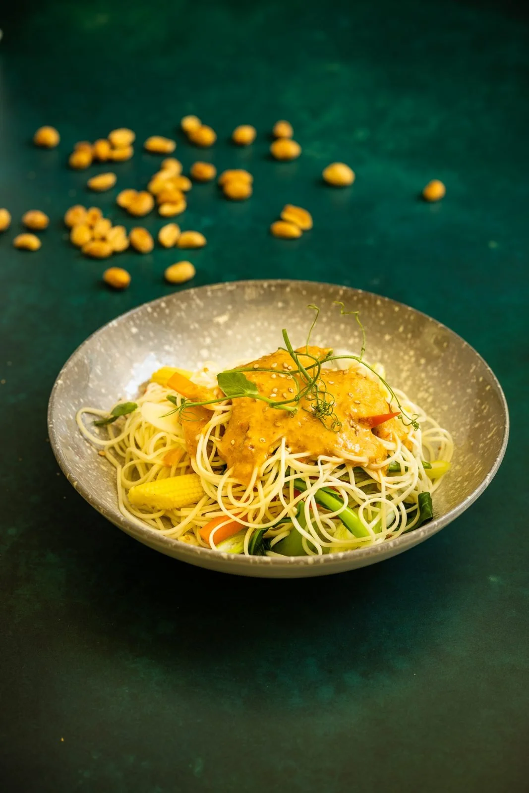 A bowl of noodle stir-fry topped with a sauce and garnished with fresh greens, on a dark green surface with scattered chickpeas in the background.