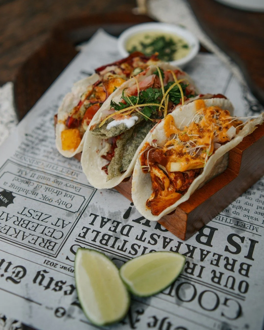 Three tacos with various fillings on a wooden tray, garnished with cilantro, accompanied by lime wedges and a side of dipping sauce, resting on newspaper.