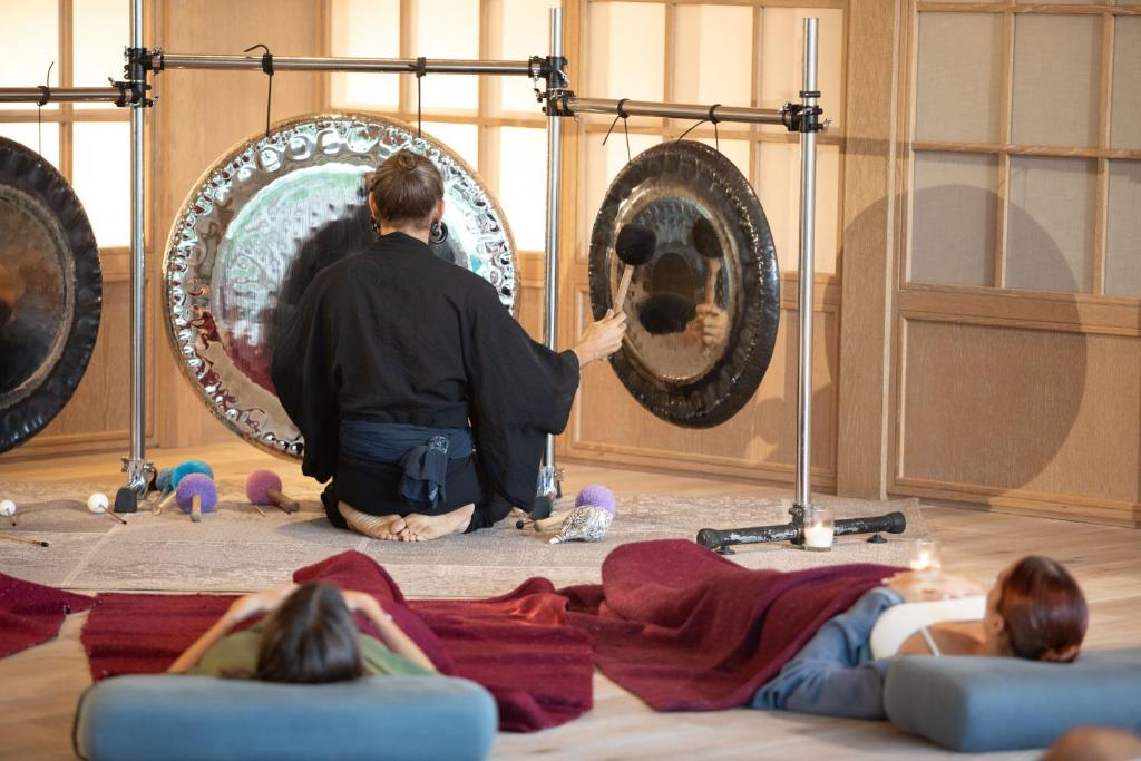 Someone playing large gongs with mallets while two people lie on mats in front of them, possibly participating in a sound healing session or meditation.