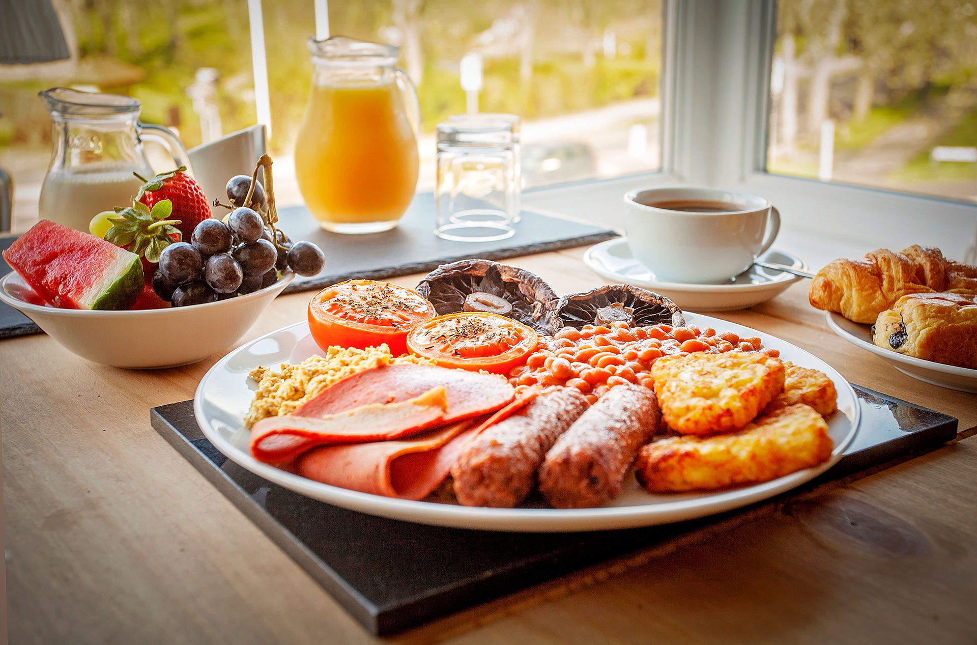 A breakfast spread on a wooden table near a window, featuring a large plate with ham, sausage links, roasted tomatoes, baked beans, scrambled eggs, and potato hash browns. Additional items include a bowl of mixed fruit, a carafe of orange juice, a glass of water, a cup of coffee, and croissants.