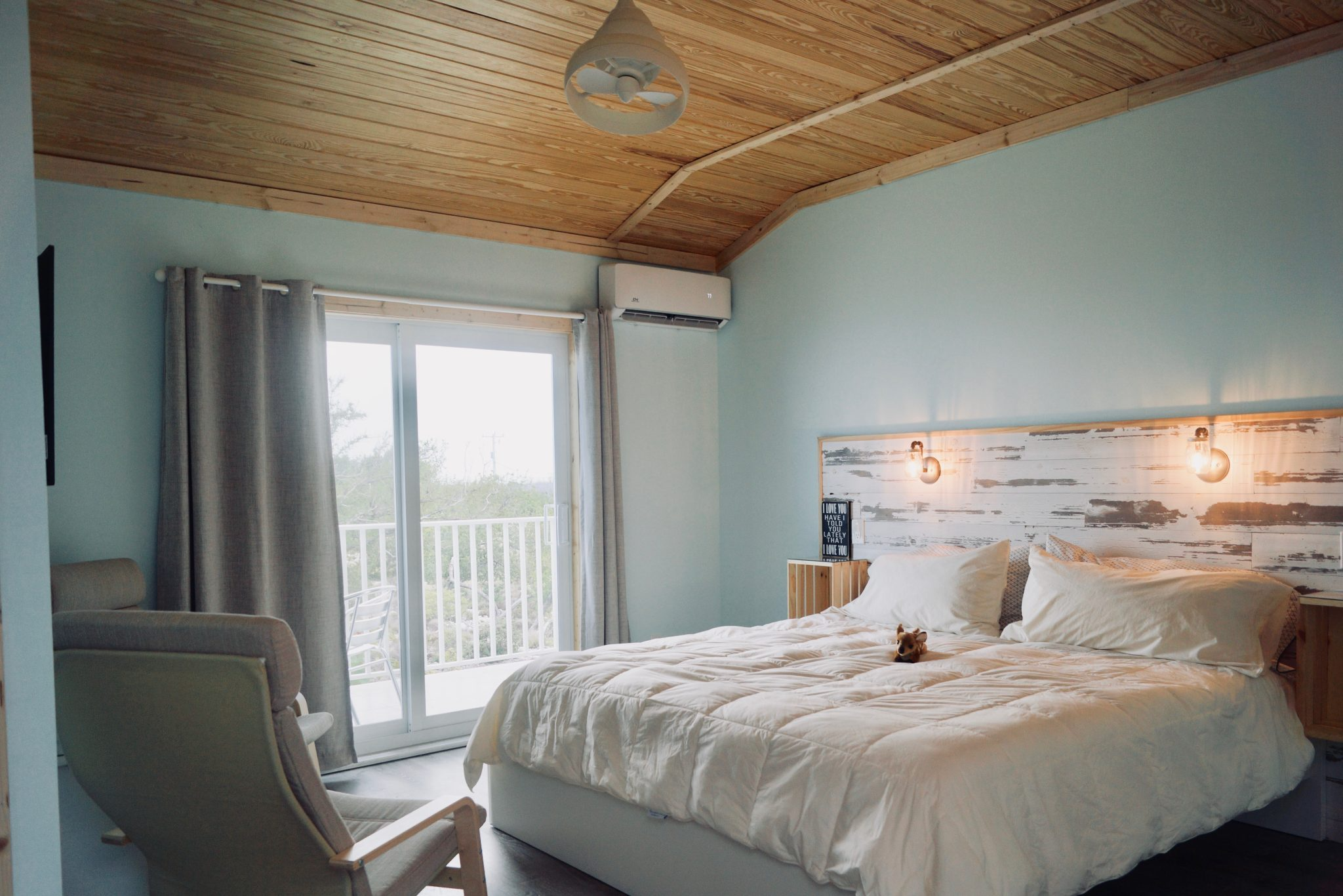 A cozy bedroom with a wooden ceiling, sliding glass door leading to a balcony, and a large bed with white bedding and pillows. There is a small stuffed animal on the bed and two reading lights attached to a distressed wood headboard. An armchair is visible near the door and a white curtain hangs over the door.