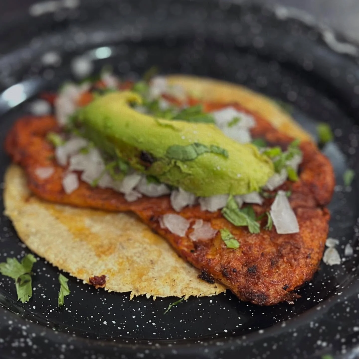 Close-up of a Mexican street taco featuring grilled chicken, chopped onions, cilantro, a slice of avocado, and a soft corn tortilla in a black speckled plate.