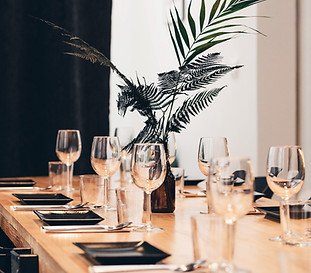 Dining table set with wine glasses, black plates, and a vase with a large fern and decorative grass.