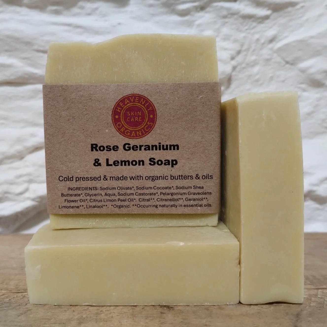 Handmade soap labeled Rose Geranium & Lemon. The soap is pale yellow and rectangular, sitting on a wooden surface with a textured white background.