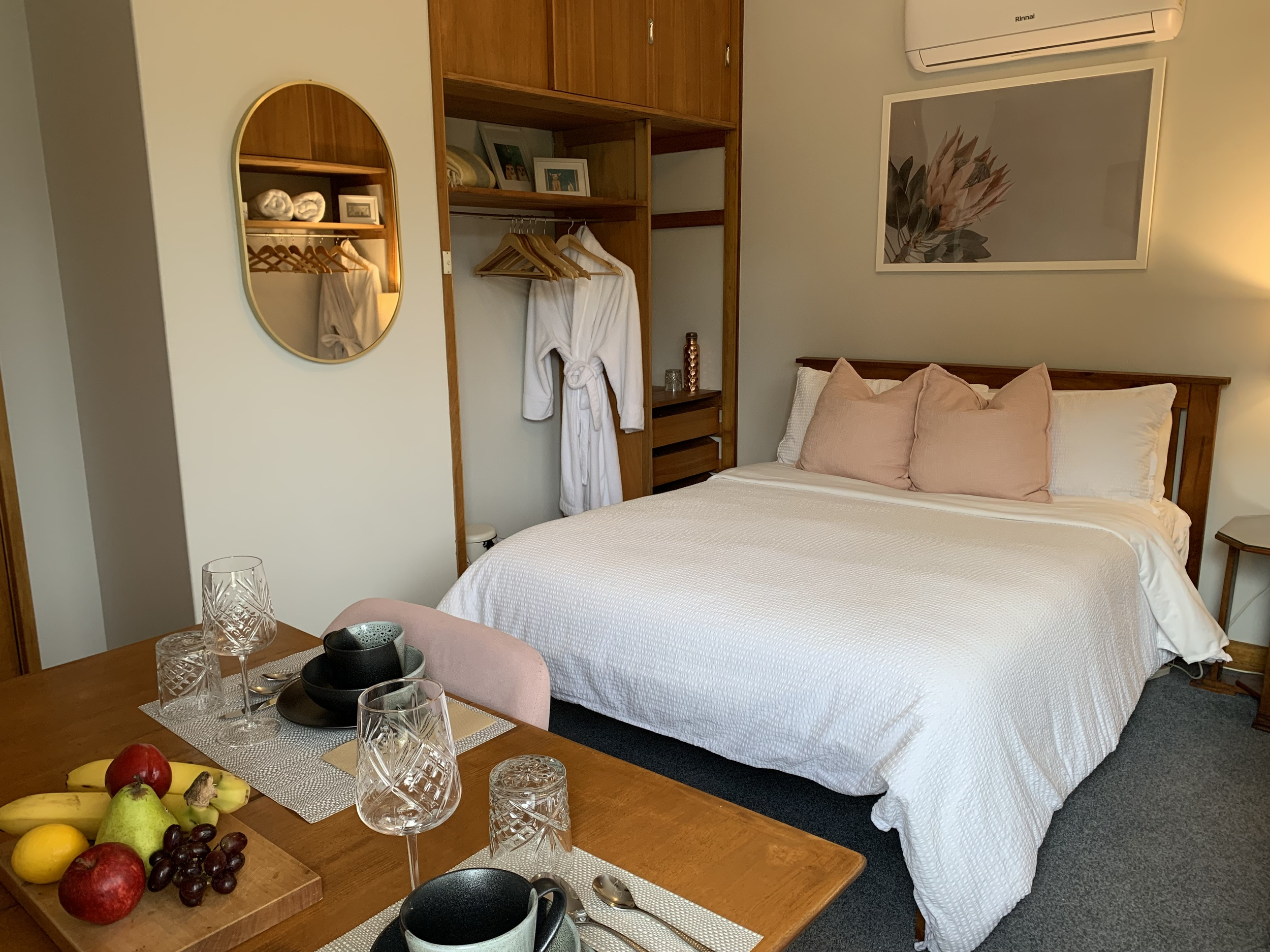 A bedroom with a bed, wooden headboard, and bedside table. There is a wooden wardrobe with open shelves and a robe hanging. A framed floral art piece is on the wall, and an air conditioning unit is visible. A table in the foreground is set with glasses, bowls, and a fruit platter with apples, bananas, pears, and grapes.