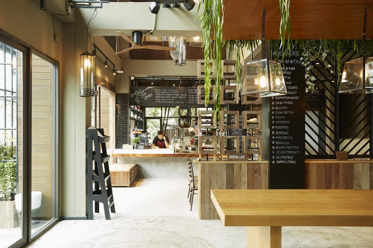 Interior of a modern cafe with wooden accents, black chalkboard menu, hanging light fixtures, and greenery. A counter with a barista preparing drinks is visible in the background.