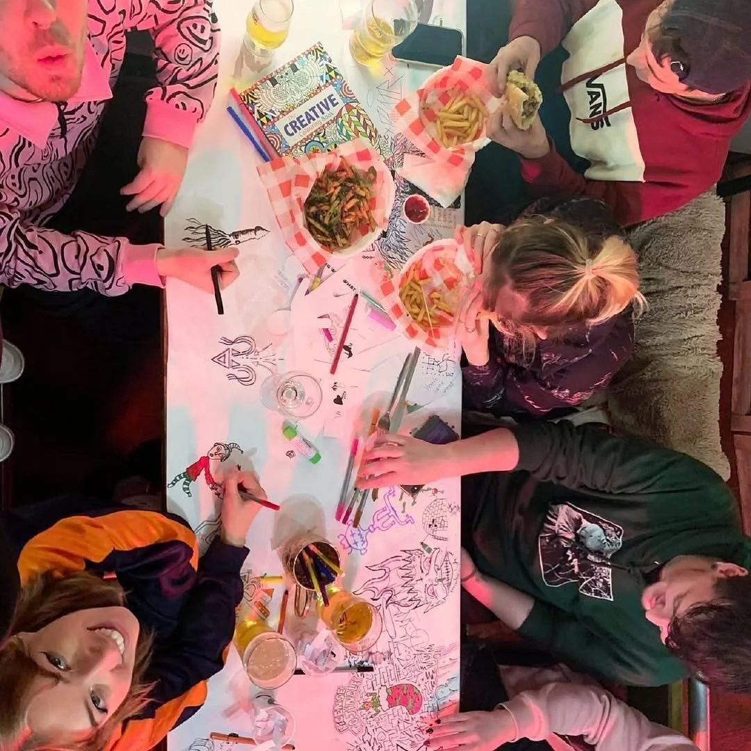 A group of people sitting around a table filled with food, drinks, art supplies, and coloring pages. Some are eating fries and sushi, others are drawing and coloring.